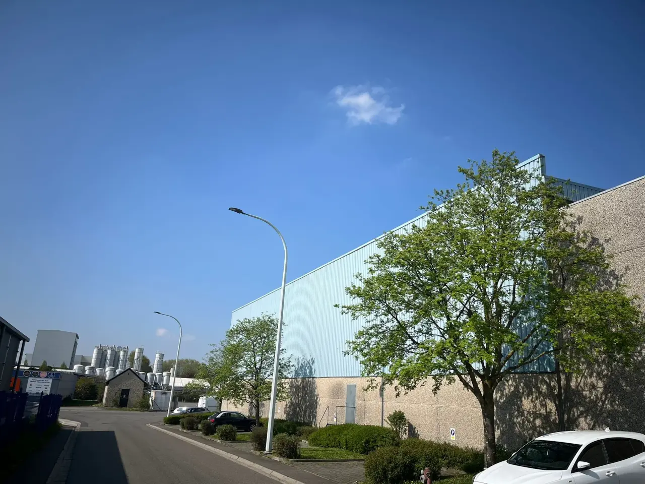 Industriegebäude mit Metallverkleidung, Parkplatz und Grünflächen unter blauem Himmel.