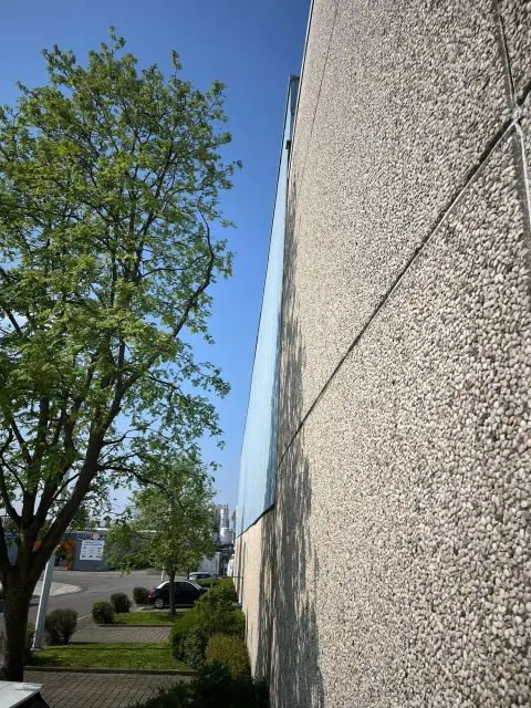 Modernes Gebäude mit Kieselfassade neben grünem Baum und Parkplatz unter blauem Himmel.