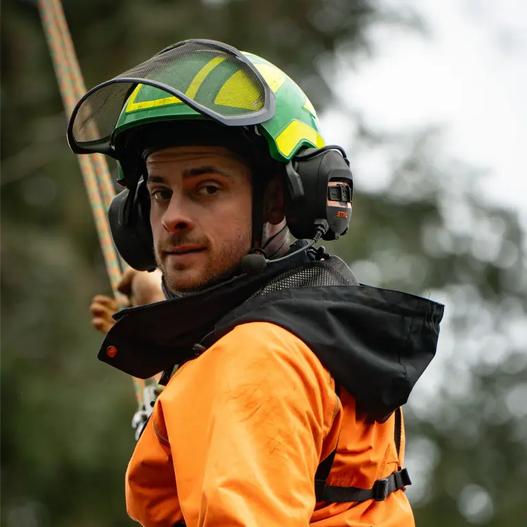 Mann mit Schutzausrüstung beim Klettern oder Abseil mit Helm, Headset und orangefarbener Jacke.