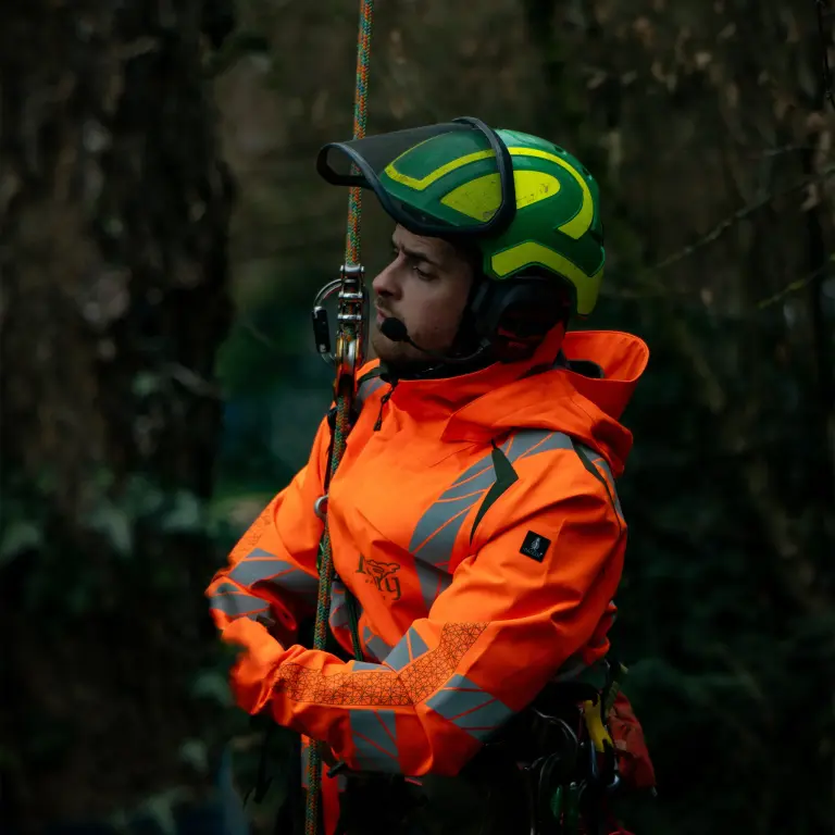 Mann in orangefarbener Jacke und grünem Helm beim Klettern an einem Seil in einem Wald.